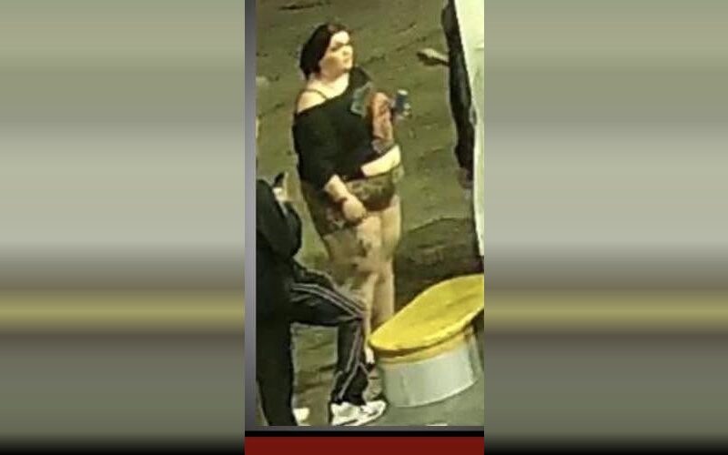 Woman standing outdoors at night near a yellow-topped bin, holding a drink.