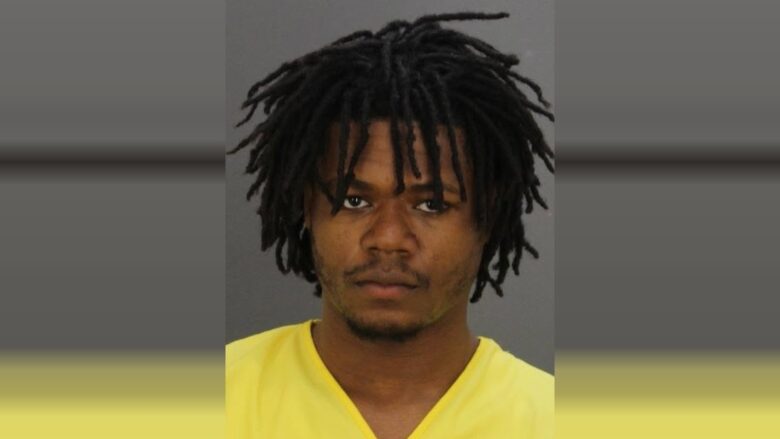 Mugshot-style portrait of a man in a yellow shirt against a gray background.