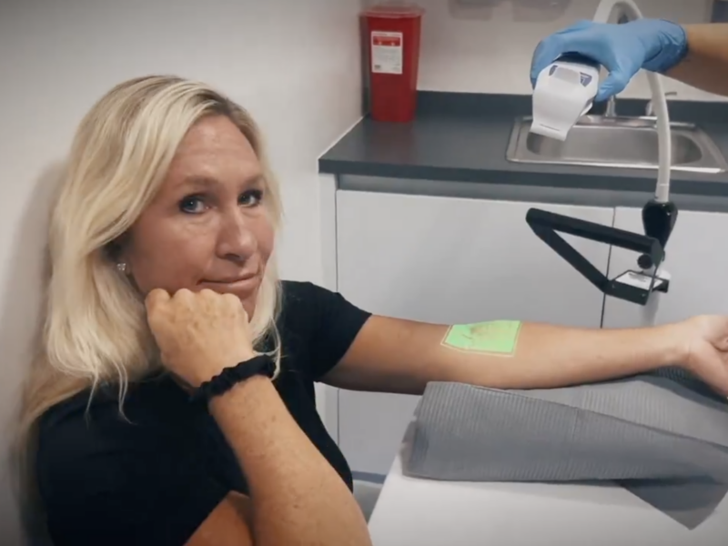 Woman receiving a medical procedure with her arm extended in a clinical exam room.