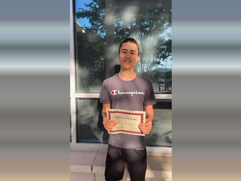 Young man standing outdoors holding a certificate in front of a reflective glass window.