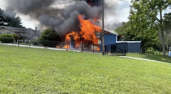 Blue outbuilding engulfed in flames with thick smoke rising over a green lawn.