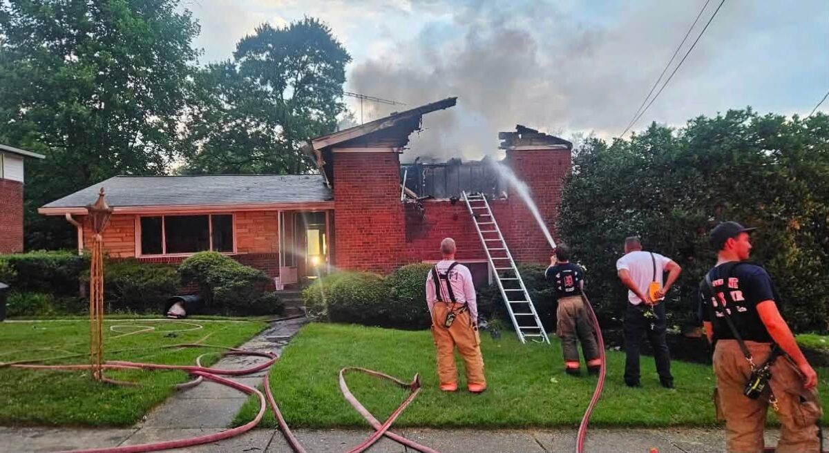 Firefighters spray water on a burning brick house with smoke rising from the roof.