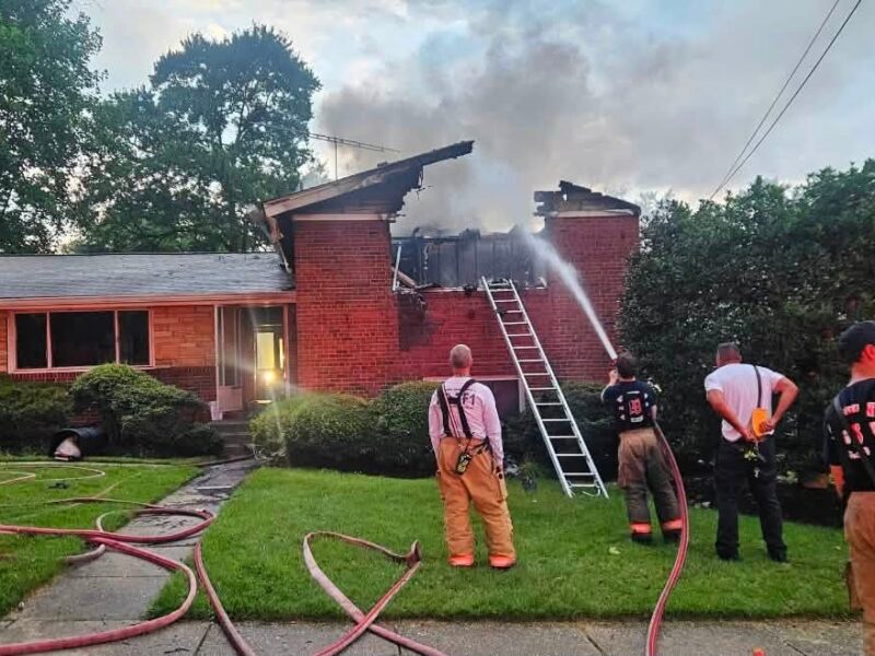Firefighters spray water on a burning brick house with smoke rising from the roof.