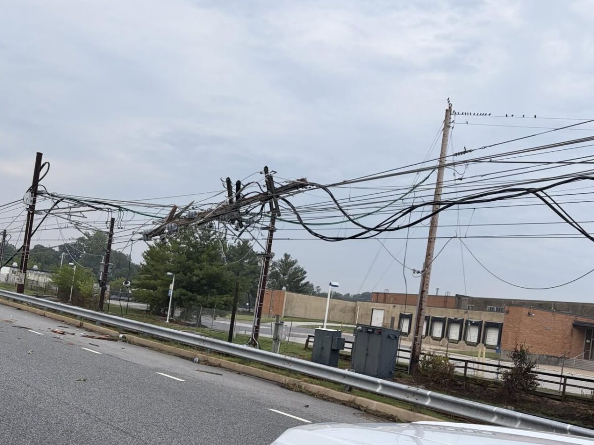 Sagging utility wires and poles line a roadside beside a brick building under a cloudy sky.