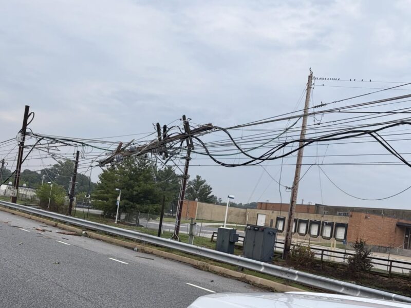Sagging utility wires and poles line a roadside beside a brick building under a cloudy sky.