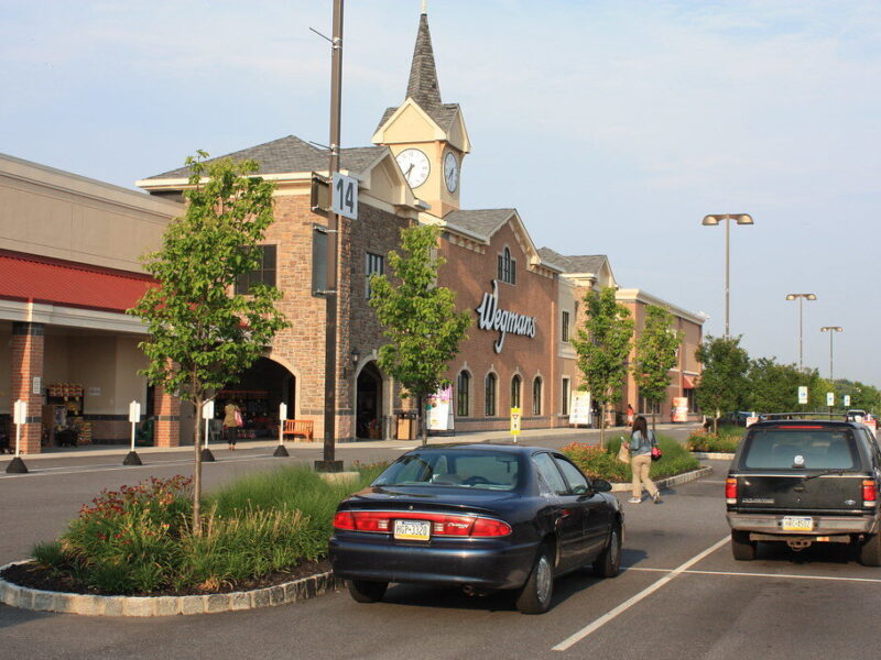 Wegmans storefront in a suburban shopping center with parked cars and a clock tower.