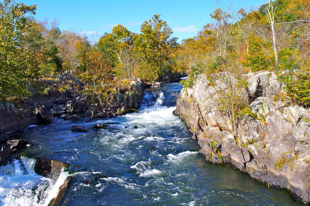 Rushing river waterfall between rocky cliffs with autumn trees under a clear blue sky.