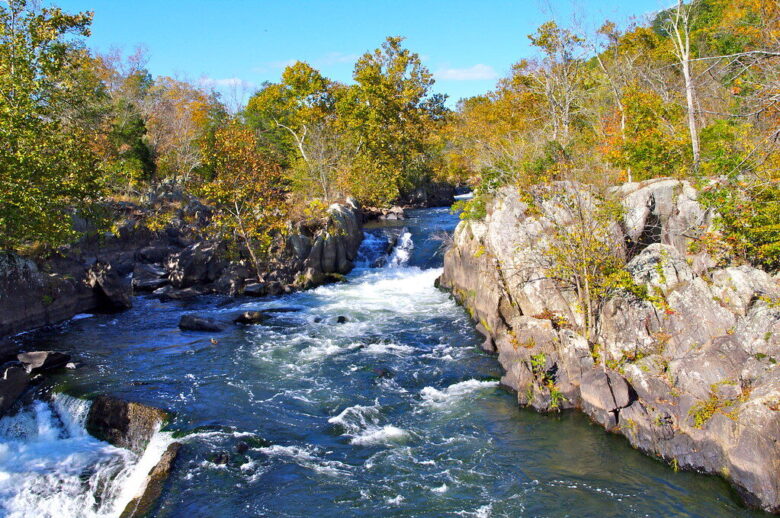 Rushing river waterfall between rocky cliffs with autumn trees under a clear blue sky.