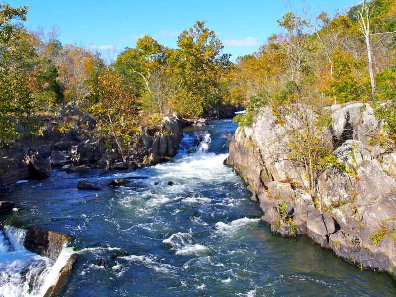 Rushing river waterfall between rocky cliffs with autumn trees under a clear blue sky.