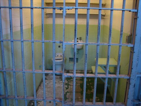 Small prison cell with barred door, a sink, toilet, and worn green walls.