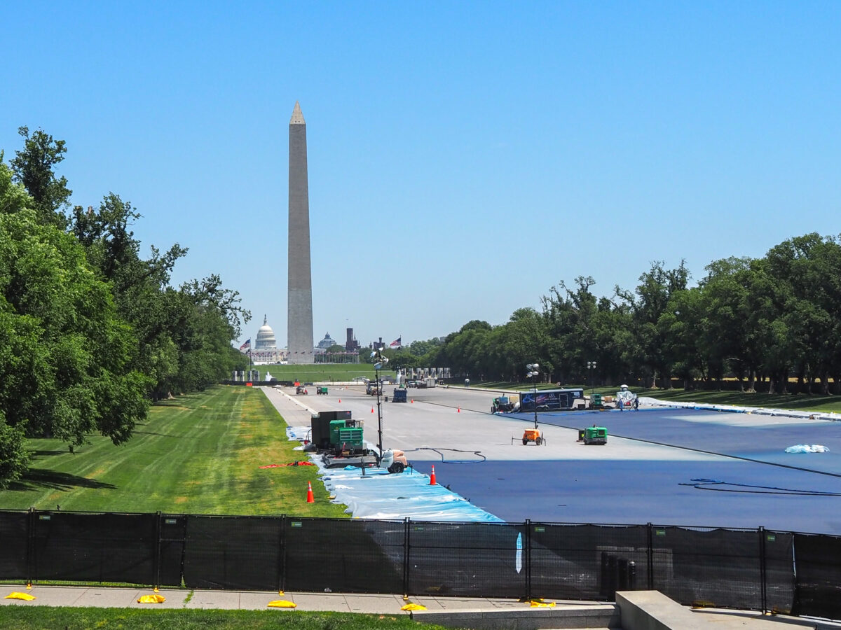 Washington Monument rises beyond a large construction site along the National Mall under a clear blue sky.