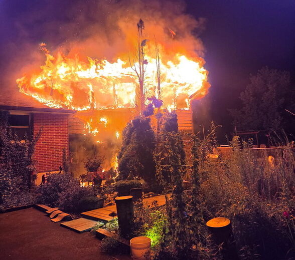 House roof engulfed in flames at night with firelighting the yard and smoke above.