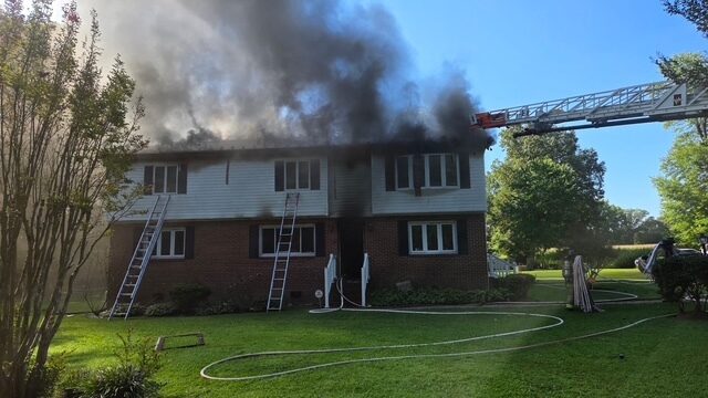 Firefighters battle heavy smoke pouring from a house as ladders reach the roof and hoses line the lawn.