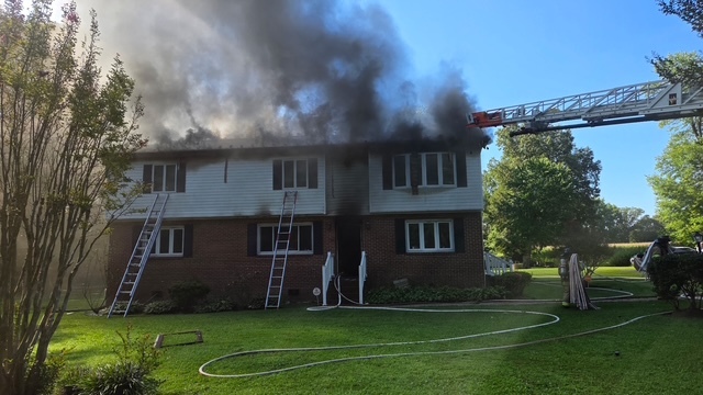 Firefighters battle heavy smoke pouring from a house as ladders reach the roof and hoses line the lawn.