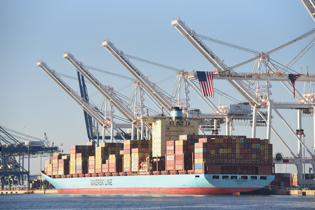 Container ship docked at an industrial port with towering cargo cranes and stacked shipping containers.