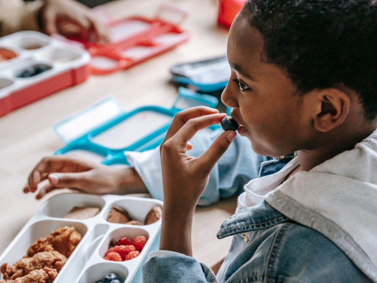 Crop African American child in casual clothes sitting at table with plastic lunch box while having snack