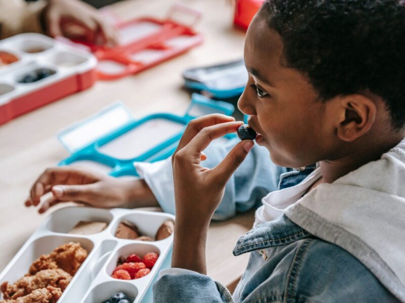 Crop African American child in casual clothes sitting at table with plastic lunch box while having snack